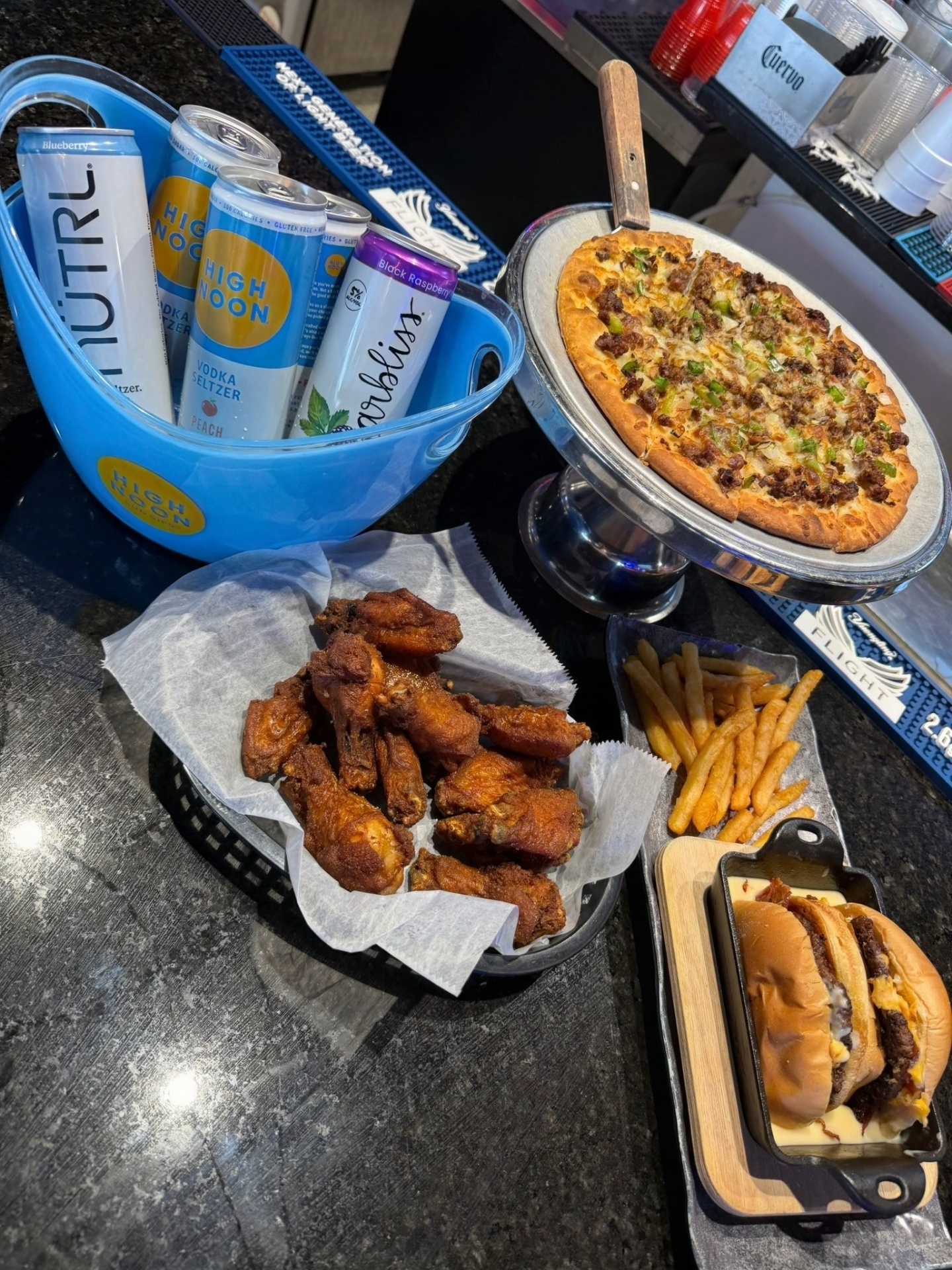 Bar top spread with a bucket of High Noon and Nutrl seltzers, a sausage and pepper pizza on a pedestal, jumbo dry-rub wings, fries and a smashburger