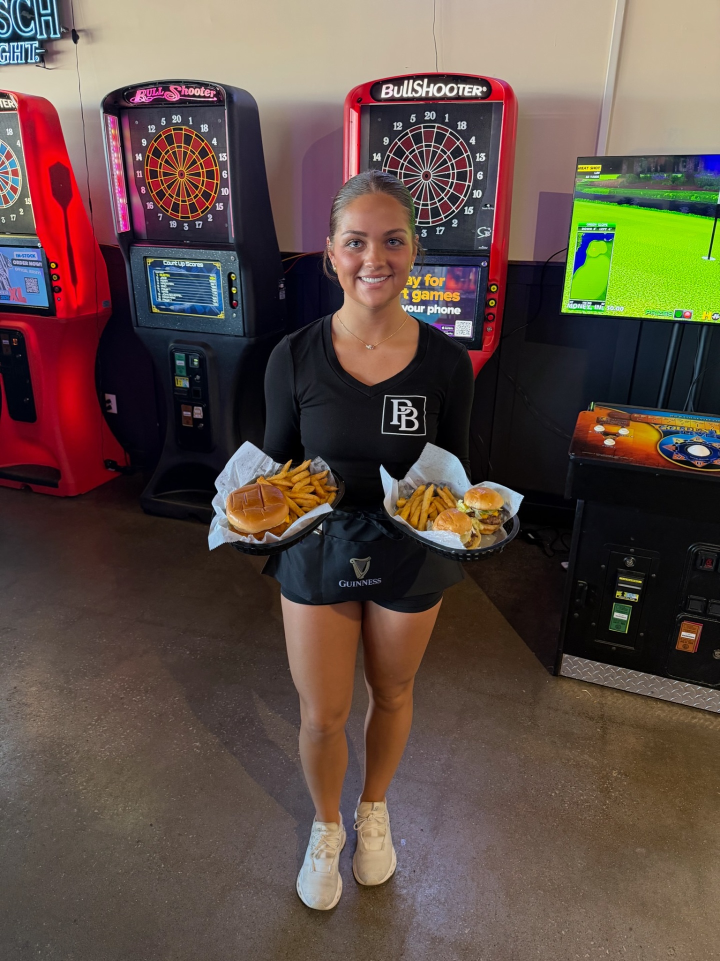 Penalty Box server in front of the BullShooter dart machines and golf game holding two burger and fries baskets