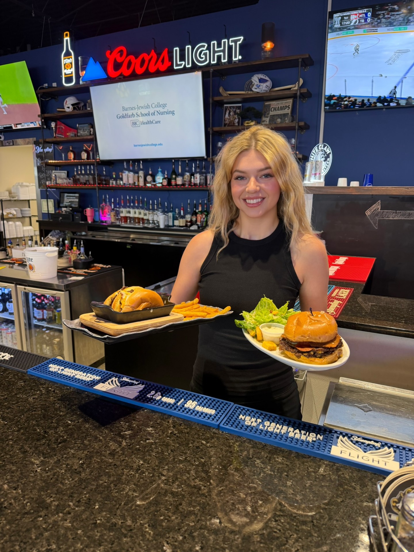 Penalty Box server at the bar under the Coors Light neon holding two burger plates with seasoned fries and a Caesar salad