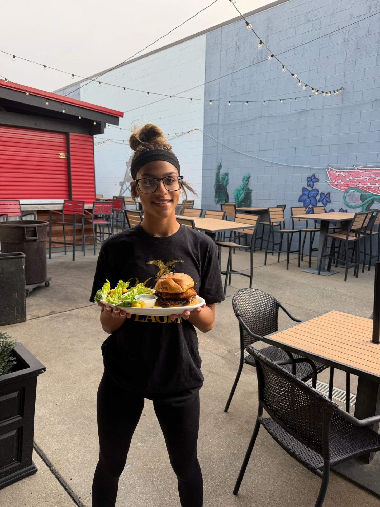Penalty Box server on the patio holding a crispy chicken sandwich with a Caesar salad in front of the mural