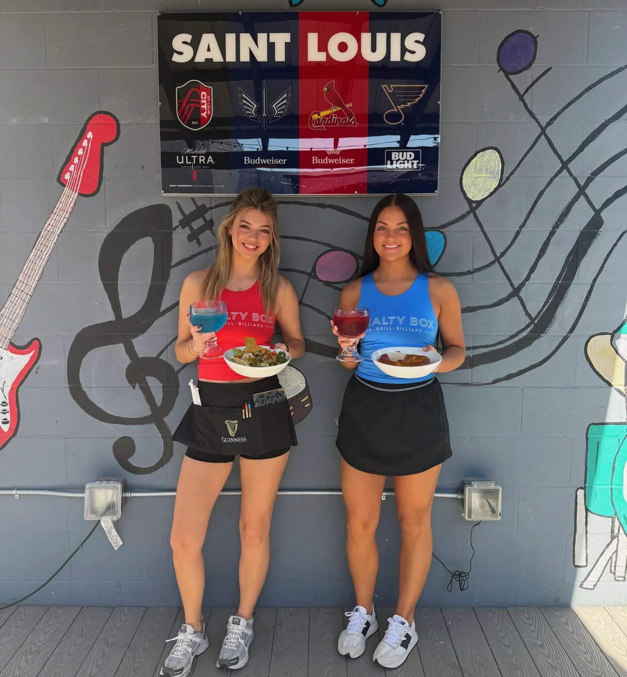Two Penalty Box servers in front of the Saint Louis sports mural with fishbowl margaritas, taco salad and bavarian pretzel