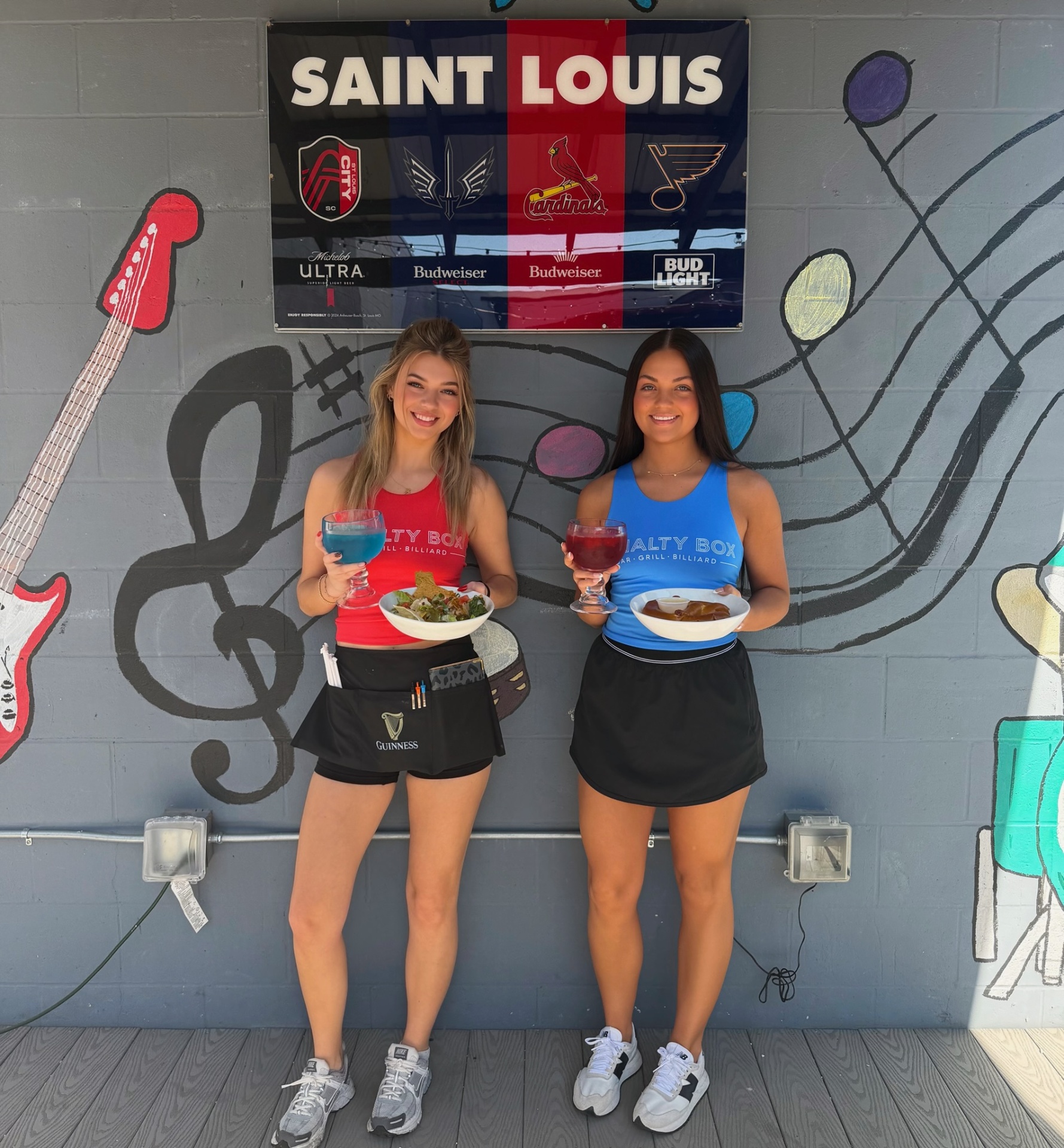 Two Penalty Box servers in front of the Saint Louis sports mural with fishbowl margaritas, taco salad and bavarian pretzel